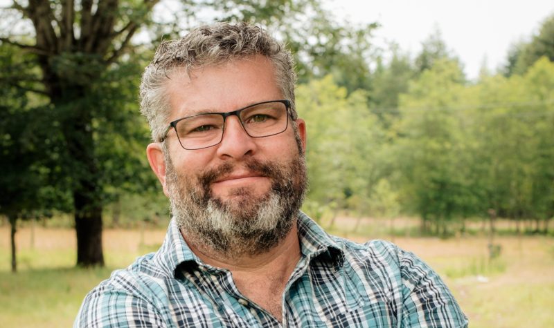 A bearded man with glasses, grey hair and a plaid shirt stands in a grassy area with trees in the background.