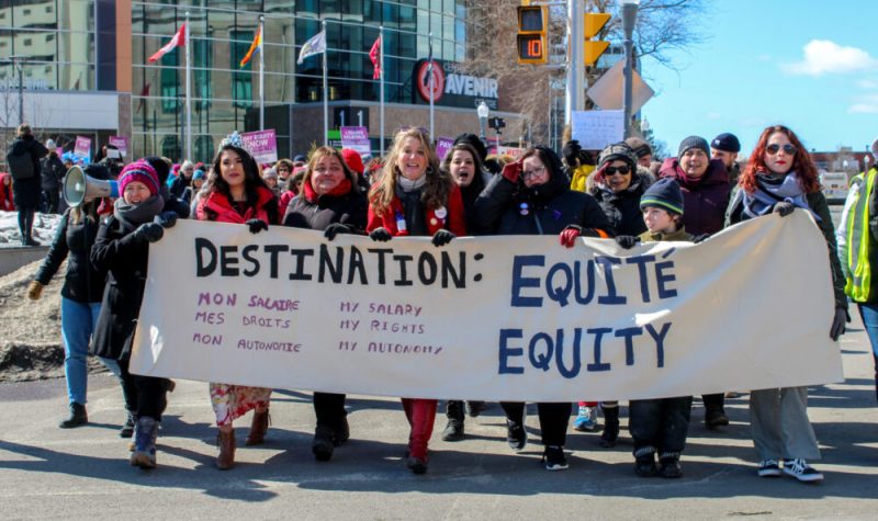 Groupe de gens qui marchent ensemble dehors au Centre Avenir avec une affiche qui lit: DESTINATION: mon salaire, mes droits, mon autonomie ÉQUITÉ / my salary, my rights, my autonomy EQUITY