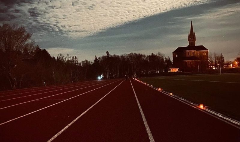 Des marcheuses avec des lampes sur la piste d'athlétisme bornée de bougie à l'Université Sainte-Anne sur une soirée avec un seulement un peu de nuages. En arrière plan, on voit l'Église Sainte-Marie, la plus grande église en bois dans l'Amérique du Nord. Le ceil paraît illuminé par la lumière de la lune.