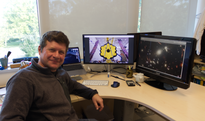 Marcin Sawicki est assis devant son bureau. Derrière lui, sur ses deux écrans d'ordinateur, on voit le James Webb Telescope et une photo de l'espace