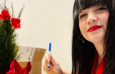 Dame cheveux noirs portant une chemise rouge et tenant un crayon en train de signer des cartes de Noël.