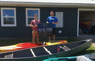 Canoes and kayaks in foreground, then two young men holding a sign that reads Maritime Paddle, with a green building behind them.