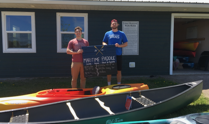 Canoes and kayaks in foreground, then two young men holding a sign that reads Maritime Paddle, with a green building behind them.
