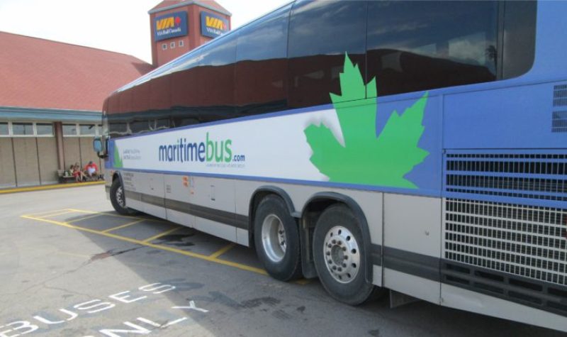 A bus with a logo for Maritime Bus on the side, featuring a large green maple leaf, parking alongside road markings reading BUSES ONLY, and in front of a VIA Rail building.