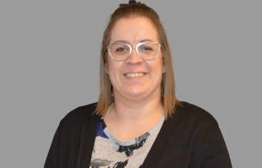 Marylise Bouchard souriante, portant des lunettes argentées, vêtues d'un cardigan noir et d'une blouse fleurit. La photo est prise sur fond gris.