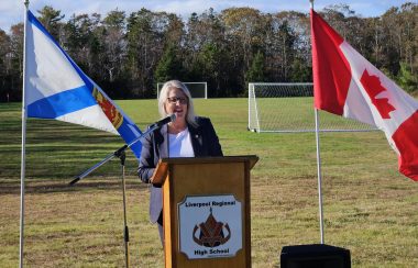 A woman stands behind a podium. The podium is outside in front of a soccer field and is flanked on either side by the flags of Nova Scotia and Canada.
