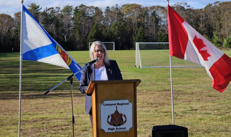 A woman stands behind a podium. The podium is outside in front of a soccer field and is flanked on either side by the flags of Nova Scotia and Canada.