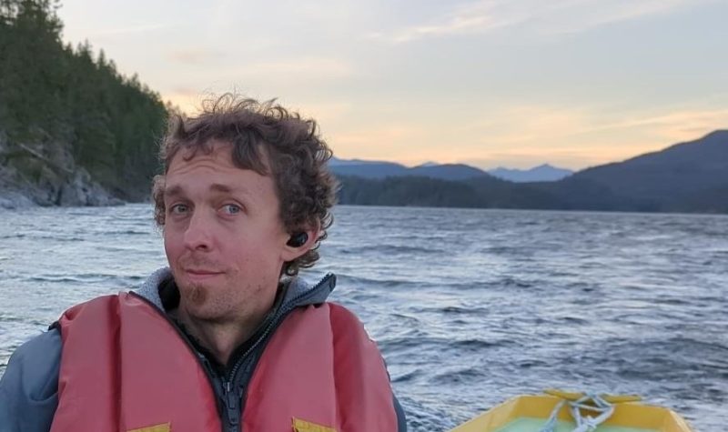 Man smiles on a boat with nature-filled background.