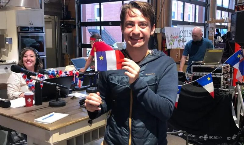Maxime Renoux, dans le centre de l'image, tenant un petit drapeau de l'Acadie dans un bureau rempli de drapeaux acadiens.