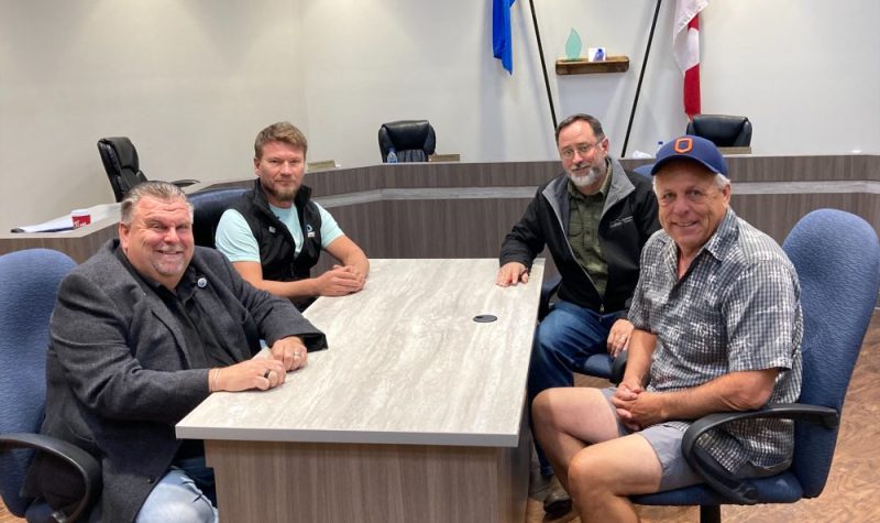 Quatre hommes sont assis autour d'une table et sourient à la caméra. La table est dans le bureau du conseil municipal. Sur le mur, il y a le drapeau canadien et le drapeau albertain.
