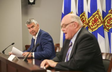 Premier Stephen McNeil and Dr. Robert Strang sit at a wooden desk at a press conference with Nova Scotia flags behind them.