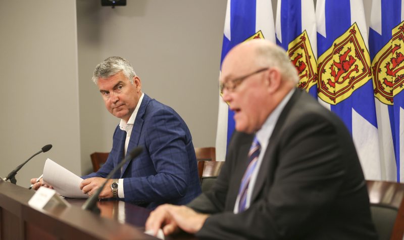 Premier Stephen McNeil and Dr. Robert Strang sit at a wooden desk at a press conference with Nova Scotia flags behind them.
