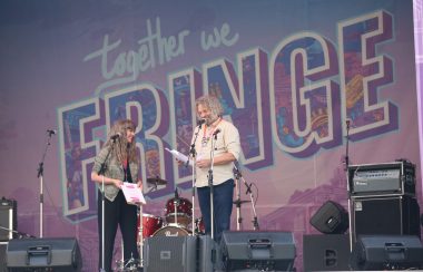 Scène de spectacle avec une affiche derrière indiquant Together We Fringe en couleur rose, violet et bleu. Des hauts-parleurs et une batterie sont aperçus sur la scène