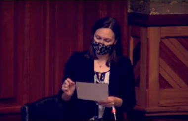 A still of Megan Mitton wearing a mask and reading from a page in legislature.