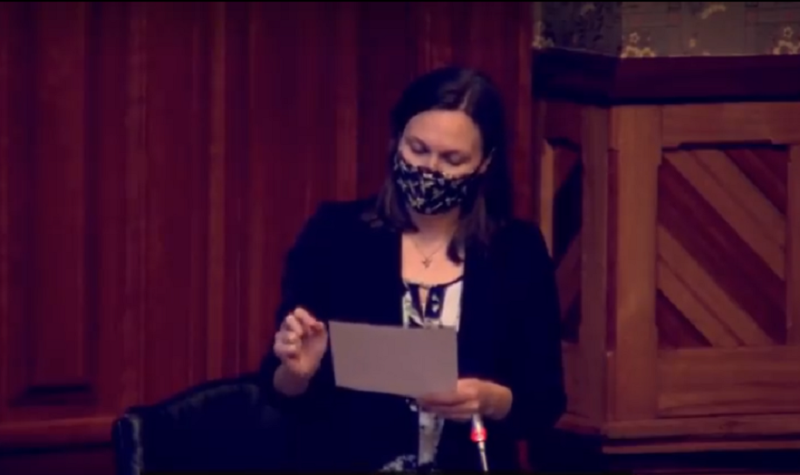 A still of Megan Mitton wearing a mask and reading from a page in legislature.