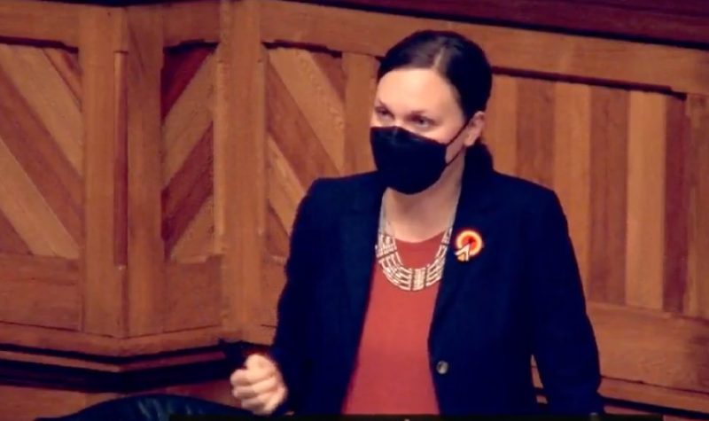 A woman with dark hair wearing a black face mask, a black jacket and a reddish-orange top with a necklace and a brooch gestures as she speaks in a wood panelled room.