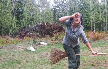 A woman in a green rocky field next to a stand of trees looks surprised as she pretends to fly away on her broom.