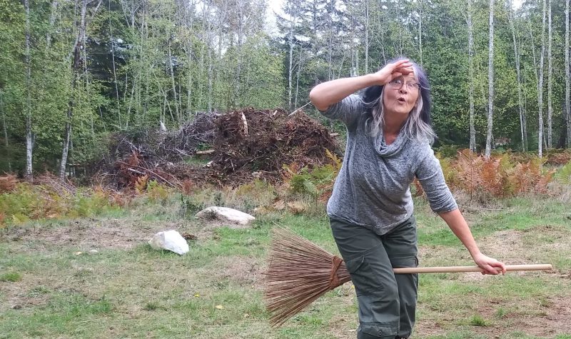 A woman in a green rocky field next to a stand of trees looks surprised as she pretends to fly away on her broom.