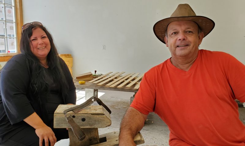 A woman and man sit in a workshop in front of a table where a canoe is being built