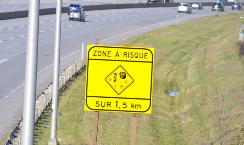 Une affiche jaune indiquant zone à risque dans 1,5 km affichant un pictogramme de vent qui souffle la neige. L'affiche est installé en bordure de l'autoroute 15