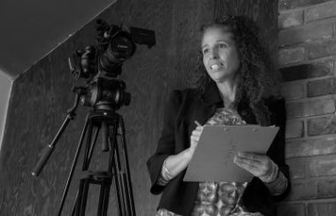 A black and white photo of Mia Golden holding a clipboard behind a camera