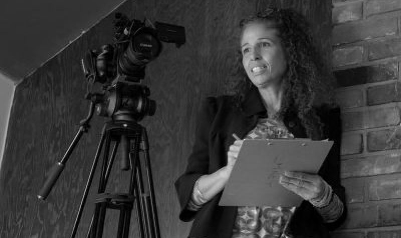 A black and white photo of Mia Golden holding a clipboard behind a camera