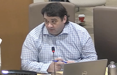 A man in a checked shirt sitting at a council table with microphone and laptop.