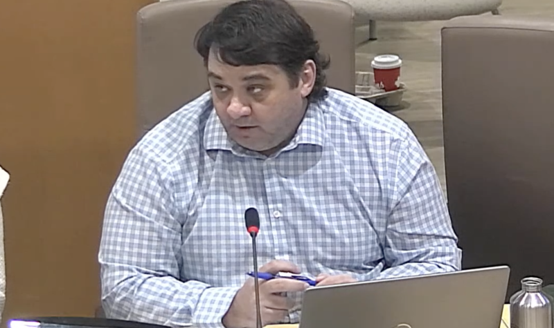 A man in a checked shirt sitting at a council table with microphone and laptop.