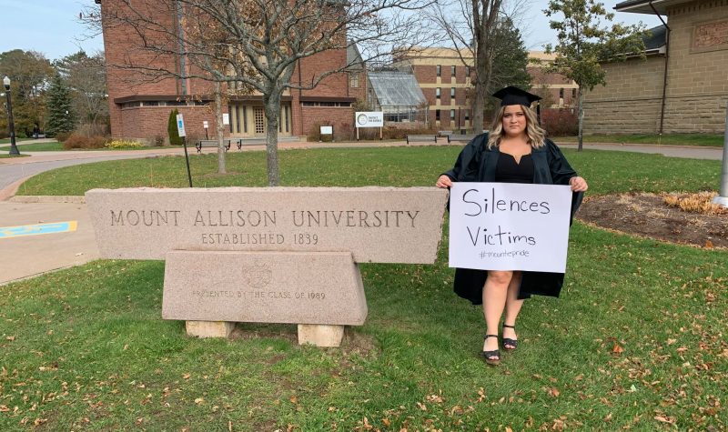 Michelle Roy en toge tenant une affiche à côté de l'enseigne de l'Université Mount Allison