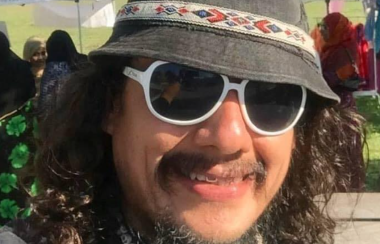 A person with long, curly hair is smiling while they wear sunglasses and a bucket hat. They are standing outdoors.