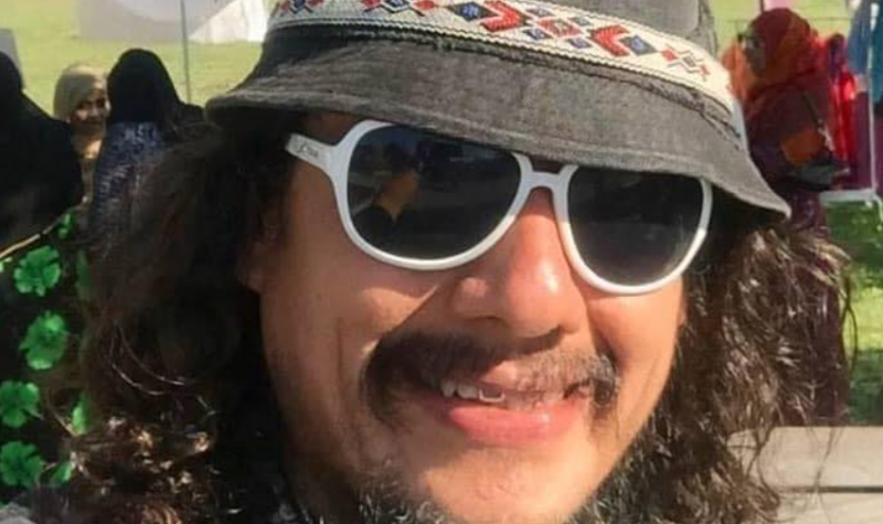 A person with long, curly hair is smiling while they wear sunglasses and a bucket hat. They are standing outdoors.