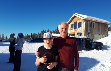 Outdoor snowy scene, one adult and child smiling at the camera, blue sky, others standing in background