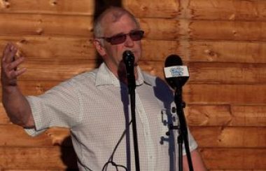 Warden candidate Michael McCrank speaks in front of two microphones wearing a white collared shirt and glasses.