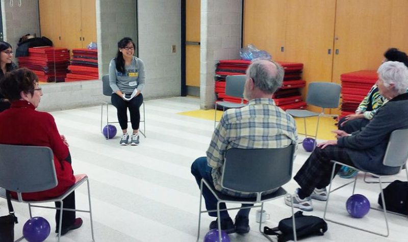 A group of seniors is seen sitting in chairs placed in a circle in a large room with exercise equipment in the background.