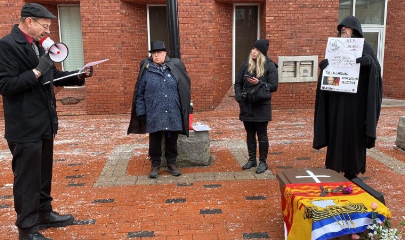 Four people, mostly wearing black, stand in a red-brick city square. One of them wears a grim reaper-style hooded robe and holds a placard. On the ground to the right is a mock-coffin fashioned from a brown plastic bin, draped in a New Brunswick flag, with a white cross and some flowers. A man on the left reads from a clipboard, speaking through a megaphone.