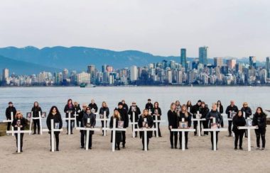 A group of family members stand with white crosses in front of them on a beach in Vancouver on a cloudy day