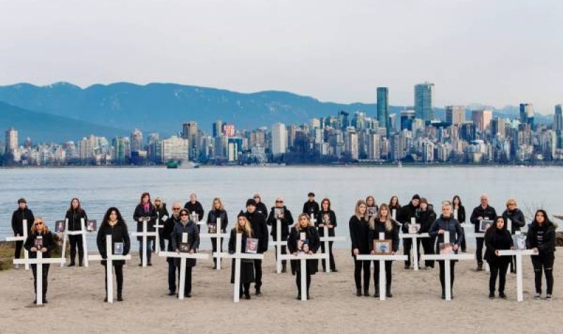 A group of family members stand with white crosses in front of them on a beach in Vancouver on a cloudy day