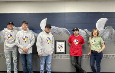 Four students wearing grey sweatshirts and a red shirt stand against a wall at school. A female teacher wearing a green shirt stands beside them.