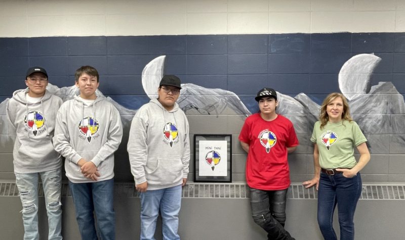 Four students wearing grey sweatshirts and a red shirt stand against a wall at school. A female teacher wearing a green shirt stands beside them.