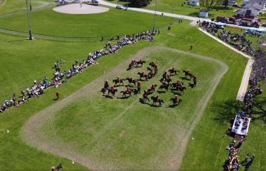 An overhead view of the RCMP Musical Ride, with more than a dozen mounties on horseback riding in four interlocking circles on a grass field.