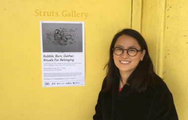 A woman wearing black stands against a yellow backdrop at Struts Gallery. There is a black and white poster on the wall.