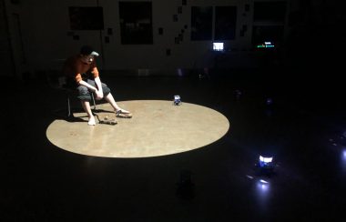 Un homme assis dans le noir éclairé par un puit de lumière, entouré de petit robots