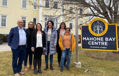 A group of people stand on a lawn beside a sign for the Mahone Bay Centre