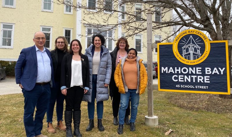 A group of people stand on a lawn beside a sign for the Mahone Bay Centre