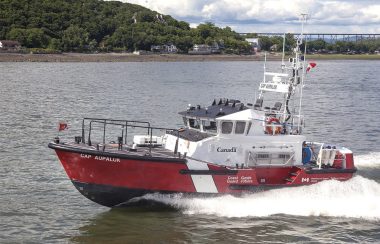 Un bateau rouge, blanc et noir navigue devant un horizon d'arbres feuillus parsemés d'habitations.