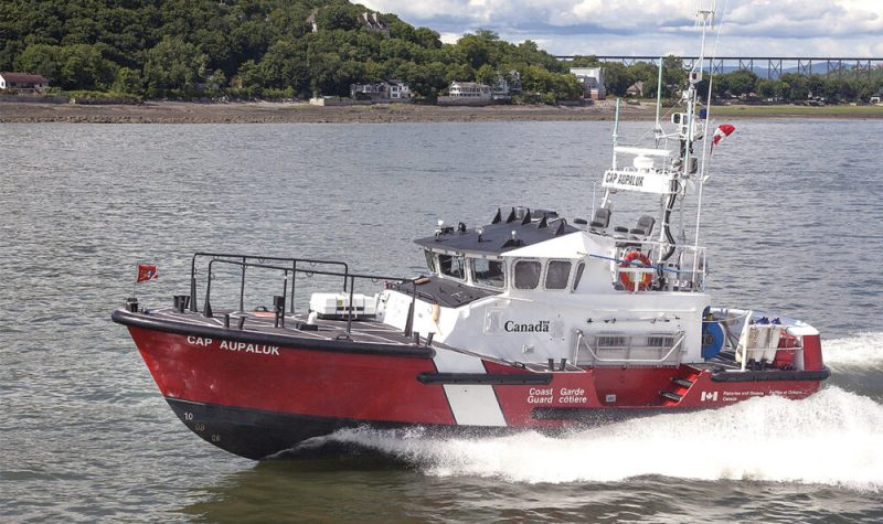 Un bateau rouge, blanc et noir navigue devant un horizon d'arbres feuillus parsemés d'habitations.