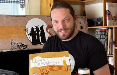 Un homme avec un chandail noir et barbe au visage avec une bande dessinée à la main portant le titre Bière dessinée dans un atelier