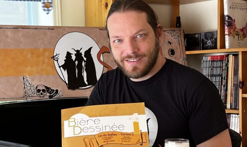 Un homme avec un chandail noir et barbe au visage avec une bande dessinée à la main portant le titre Bière dessinée dans un atelier