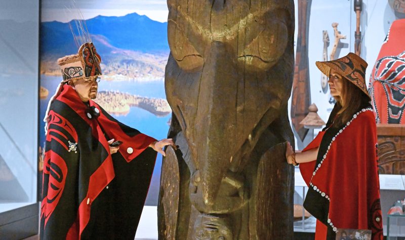 Two people dressed in traditional regalia stand on either side of a totem pole