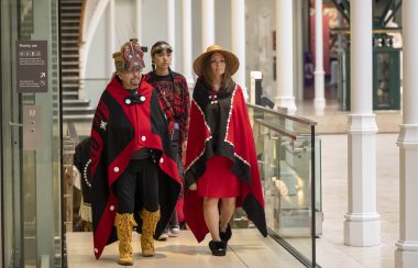 three Nisga'a representatives walk to National Museum of Scotland in a bright, airy building wearing traditional Nisga'a colours of red and black robes and garments.